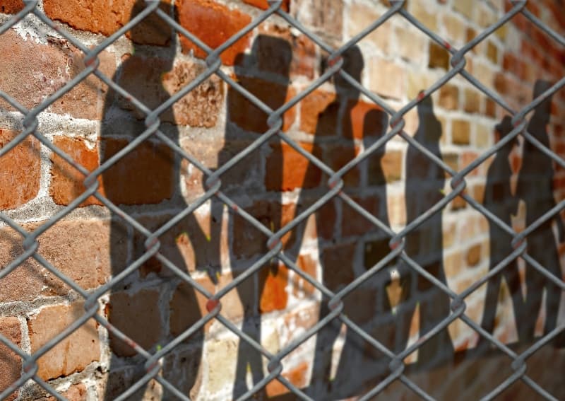 shadow outline of displaced persons on a brick wall, behind a chainlink fence